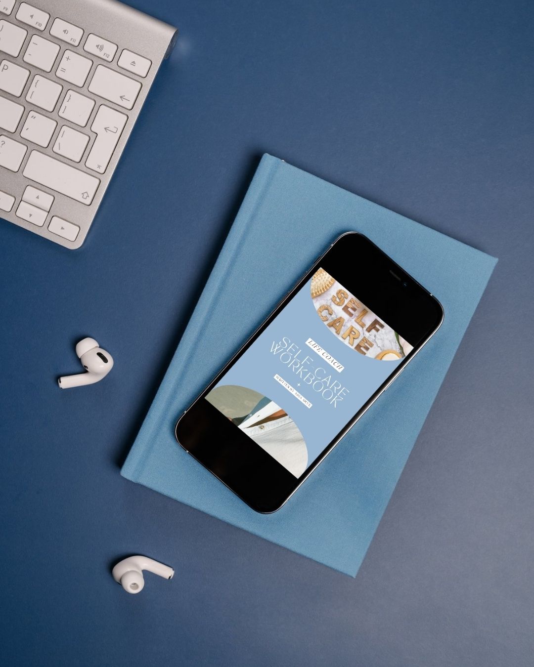 Phone with a book on a blue surface, with keyboard and earbuds nearby. Self-care Workbook.